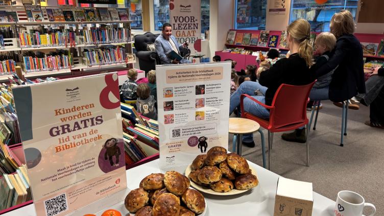Tafel met fruit en krentenbollen met op de achtergrond voorlezende wethouder met kinderen