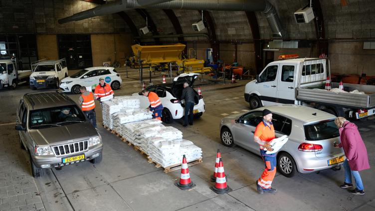 Auto’s rijden langs een uitdeelpunt waar medewerkers zakken compost klaarzetten. Inwoners stoppen kort om een zak op te halen in de grote loods.