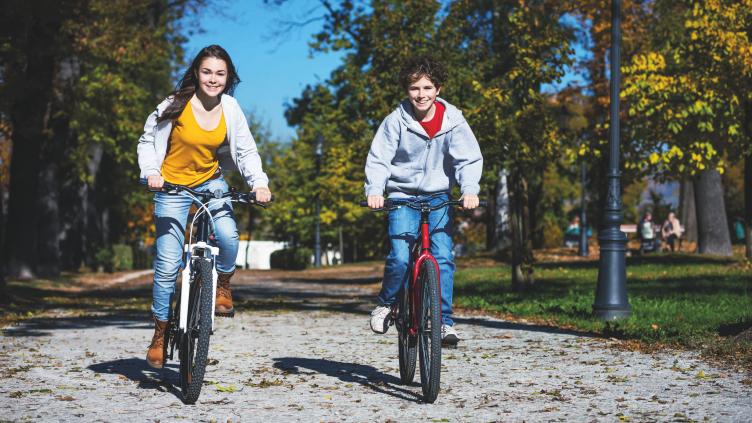 Twee kinderen fietsen naast elkaar over een onverhard pad in een groene, parkachtige omgeving met bomen en lantaarnpalen.