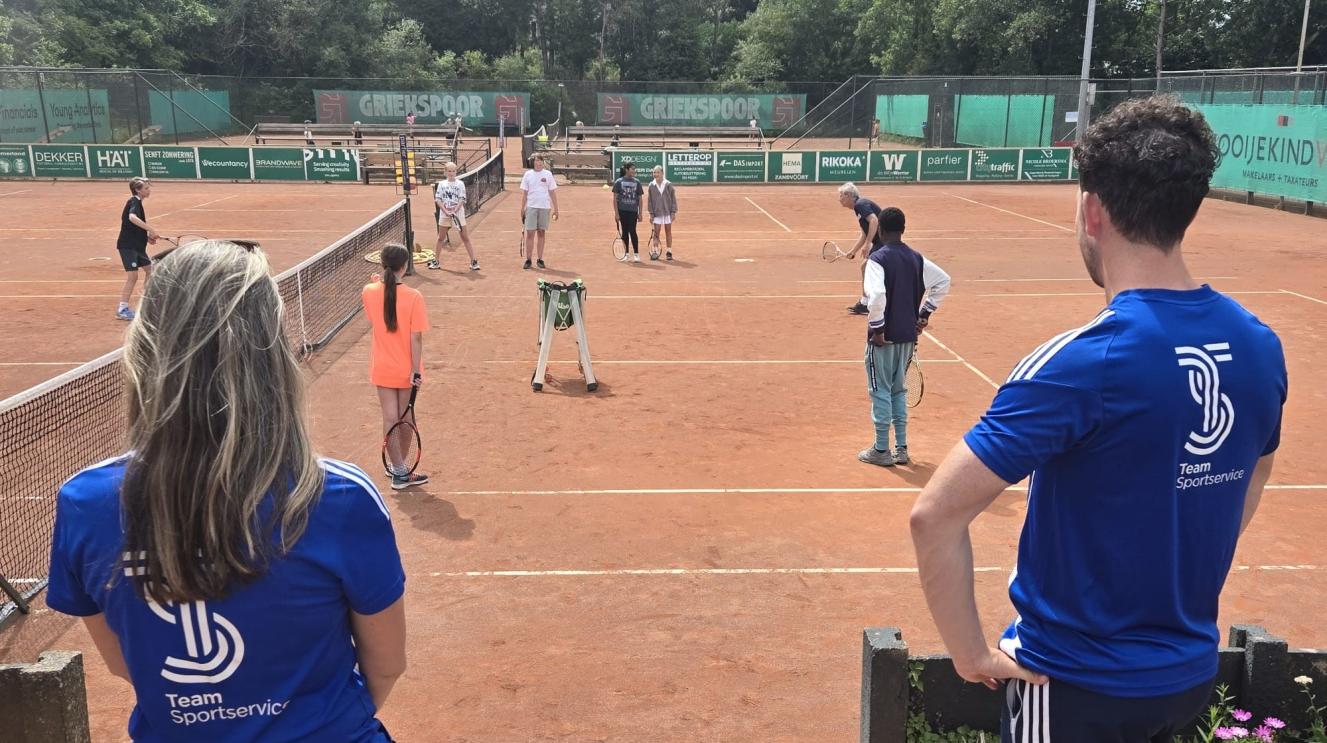 Twee medewerkers van Team Sportservice kijken naar kinderen die tennissen