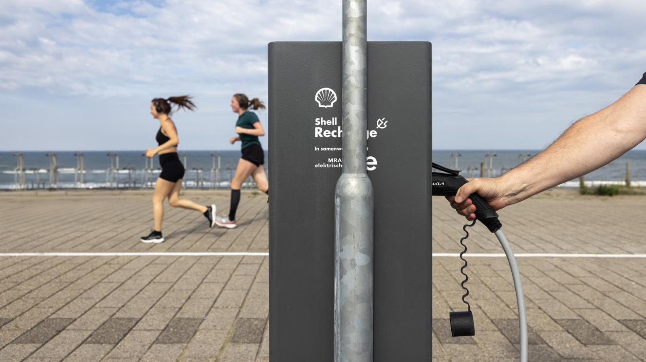 Iemand maakt gebruik van een laadpaal vlakbij het strand. In de achtergrond 2 hardlopers