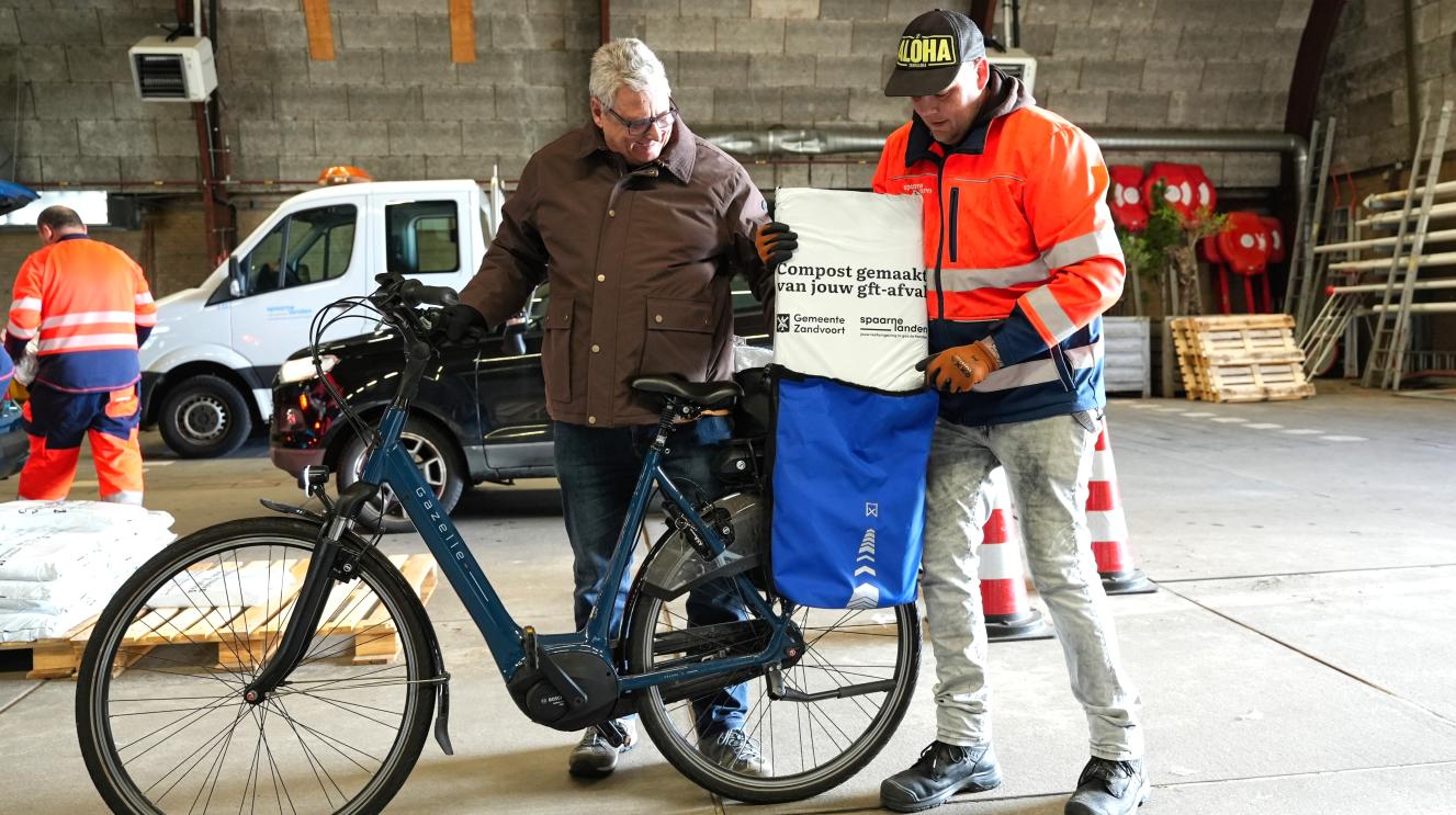 Een inwoner ontvangt een zak compost van een medewerker tijdens de uitdeeldag. Naast hen staat een fiets en op de achtergrond liggen stapels zakken compost klaar