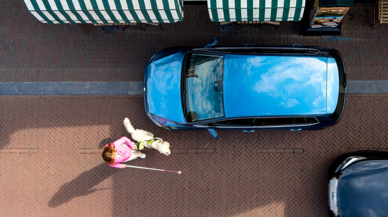 Een vrouw met een witte stok loopt langs een auto die half op de stoep geparkeerd staat